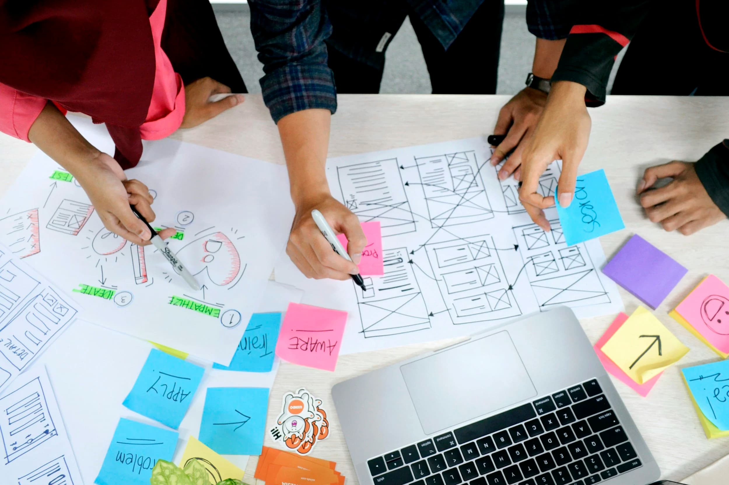 Overhead view of a design workshop: screen sketches, sticky notes and a laptop on a table.